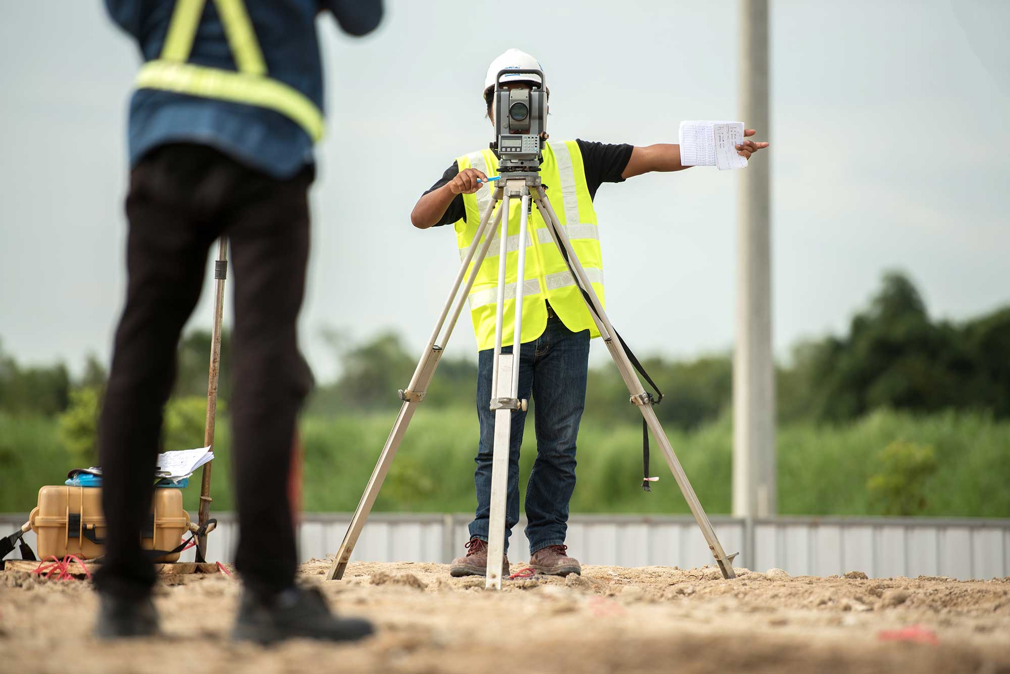 Engineers in safety vests use surveying equipment on a job site. One man is looking through the equipment and pointing off to the side.